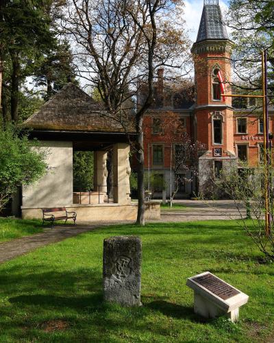 A red brick building with a corner tower and a stone gazebo in a park-like setting, featuring a visible "Rathaus" sign on the building facade.