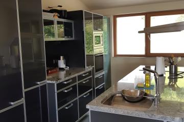 A modern kitchen interior featuring dark grey high-gloss cabinetry, granite countertops, a stainless steel sink with a bowl inside, built-in ovens, and a window looking out onto greenery.