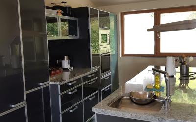A modern kitchen interior featuring dark grey high-gloss cabinetry, granite countertops, a stainless steel sink with a bowl inside, built-in ovens, and a window looking out onto greenery.
