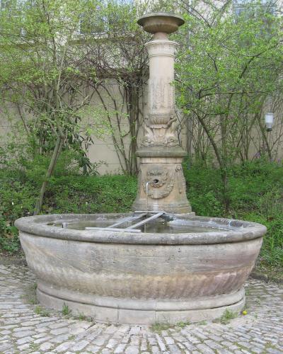 A stone fountain with a lion-head spout and a large basin, situated on a cobblestone surface with trees and a building facade in the background.