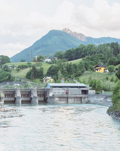A concrete hydroelectric power station and dam structure spanning a wide river, with a green forested hill and rocky outcrop in the background.