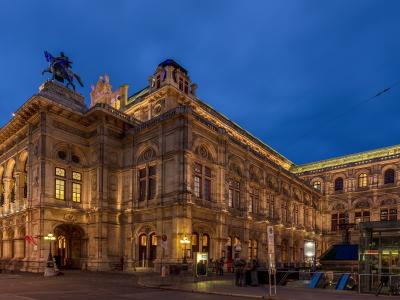 A grand, ornate historic building illuminated at dusk, featuring a prominent equestrian statue on the roof, arched windows, and a tram stop shelter in the foreground.