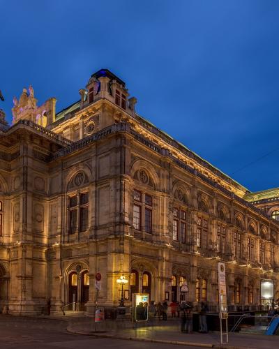 A grand, ornate historic building illuminated at dusk, featuring a prominent equestrian statue on the roof, arched windows, and a tram stop shelter in the foreground.