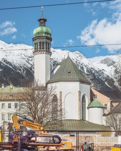 A white church with a tall tower topped by a green copper dome and a smaller octagonal chapel section, set against snow-capped mountains, with construction equipment in the foreground.