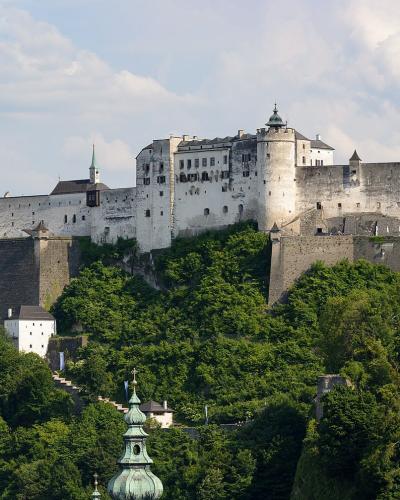 A large medieval fortress with white walls and towers sits atop a densely forested hill, with a green church spire visible in the foreground.