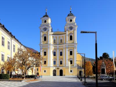 A large yellow church with two prominent towers featuring onion domes and crosses, situated in a paved town square with autumn trees and adjacent buildings under a clear blue sky.