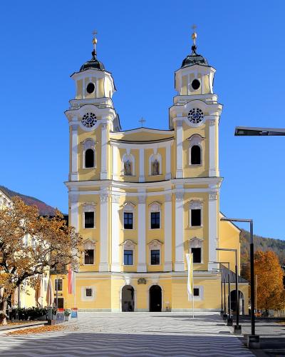 A large yellow church with two prominent towers featuring onion domes and crosses, situated in a paved town square with autumn trees and adjacent buildings under a clear blue sky.