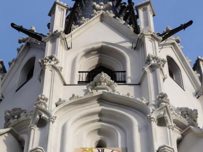 A low-angle view of the white Gothic tower of a church, featuring a clock face with golden hands, ornate stone carvings, and a dark spire topped with a golden orb and cross against a blue sky.