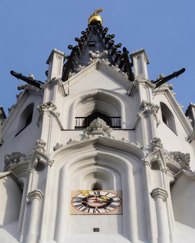 A low-angle view of the white Gothic tower of a church, featuring a clock face with golden hands, ornate stone carvings, and a dark spire topped with a golden orb and cross against a blue sky.