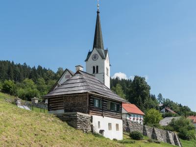 A white church with a tall, dark spire and a clock face, situated on a grassy hillside next to a wooden and stone building, under a clear blue sky.