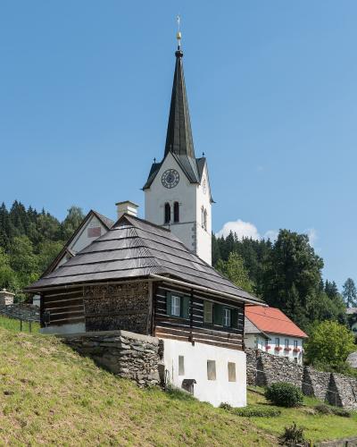 A white church with a tall, dark spire and a clock face, situated on a grassy hillside next to a wooden and stone building, under a clear blue sky.