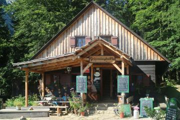 A wooden alpine hut with a sign reading "Grimminghütte" above the entrance, surrounded by green trees and outdoor seating with chalkboard menus.