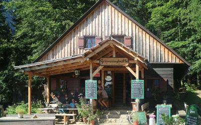 A wooden alpine hut with a sign reading "Grimminghütte" above the entrance, surrounded by green trees and outdoor seating with chalkboard menus.