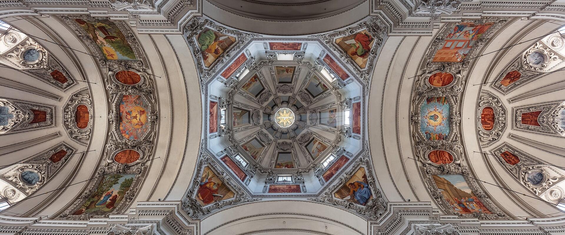 The image shows an ornate, high-ceilinged interior with a central octagonal dome, elaborate white stucco work, and numerous religious frescoes on the vaults and pendentives, characteristic of Baroque church architecture.