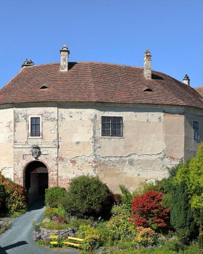 A large, historic castle complex with a prominent corner tower featuring a red-tiled roof, situated on a hillside with gardens and a clear blue sky.