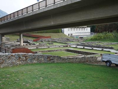The image shows the stone foundations of an archaeological site with a modern concrete overpass bridge running directly above it, a white building in the background, and a small trailer on the grass.