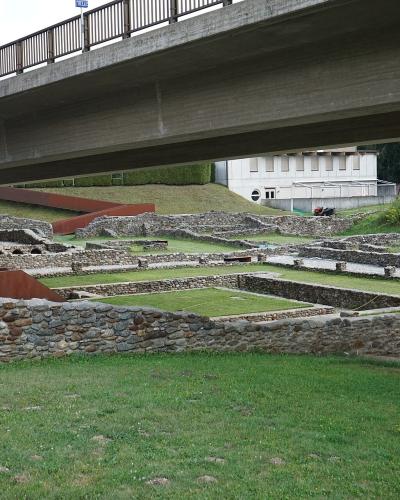 The image shows the stone foundations of an archaeological site with a modern concrete overpass bridge running directly above it, a white building in the background, and a small trailer on the grass.