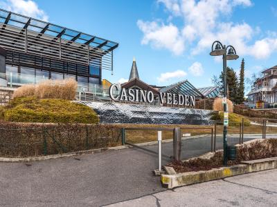 A modern building complex with a large sign reading "CASINO VELDEN" on a textured wall, set against a blue sky with clouds, featuring a glass-roofed structure and surrounding landscaping.