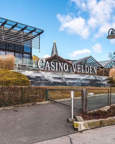 A modern building complex with a large sign reading "CASINO VELDEN" on a textured wall, set against a blue sky with clouds, featuring a glass-roofed structure and surrounding landscaping.