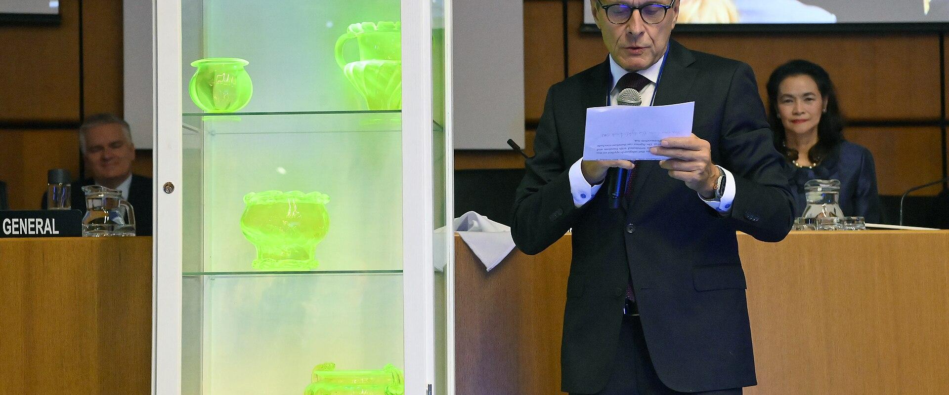 A man in a suit stands at a podium reading from a paper, next to a white display cabinet containing glowing green glass vases, inside a formal conference room with wood paneling and a "GENERAL" nameplate visible.