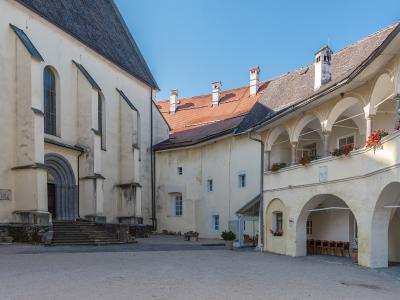 A courtyard view of a historic complex featuring a large church building on the left and a two-story arched cloister or gallery on the right, with a red-tiled roof visible in the background.