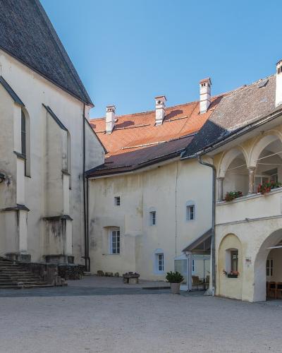 A courtyard view of a historic complex featuring a large church building on the left and a two-story arched cloister or gallery on the right, with a red-tiled roof visible in the background.