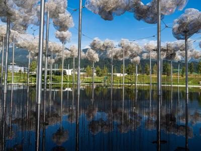 The image shows an outdoor art installation featuring numerous silver poles topped with cloud-like structures made of reflective material, situated in a pool of water with mountains in the background.