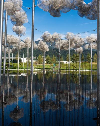 The image shows an outdoor art installation featuring numerous silver poles topped with cloud-like structures made of reflective material, situated in a pool of water with mountains in the background.