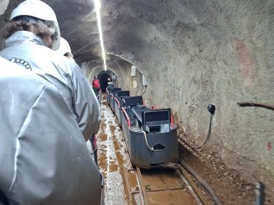 A view from inside a moving mine train carriage traveling through a narrow, concrete-lined tunnel with wet tracks and other passengers wearing white helmets and protective suits.