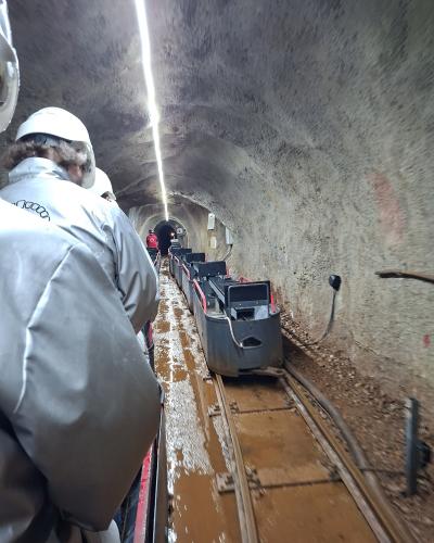 A view from inside a moving mine train carriage traveling through a narrow, concrete-lined tunnel with wet tracks and other passengers wearing white helmets and protective suits.