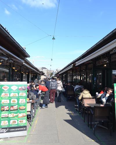 A crowded outdoor pedestrian walkway lined with long, single-story restaurant buildings featuring awnings and outdoor seating, with a green menu board for "Asia Time" visible on the left.