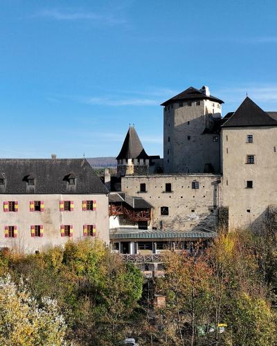 A large medieval castle complex with a prominent round tower, a rectangular keep, and a long wing featuring red and yellow shutters, set against a forested hillside under a blue sky.