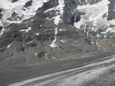 A high-angle view of a large, debris-covered glacier flowing down a steep, rocky mountain face under a blue sky with scattered clouds.