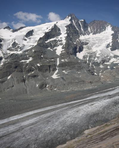 A high-angle view of a large, debris-covered glacier flowing down a steep, rocky mountain face under a blue sky with scattered clouds.