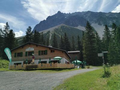 A traditional wooden alpine guesthouse (Gasthaus) with green shutters and outdoor seating, set against a backdrop of steep, rocky mountain peaks and coniferous trees.