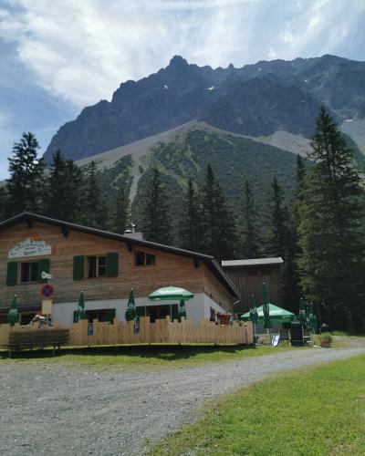 A traditional wooden alpine guesthouse (Gasthaus) with green shutters and outdoor seating, set against a backdrop of steep, rocky mountain peaks and coniferous trees.