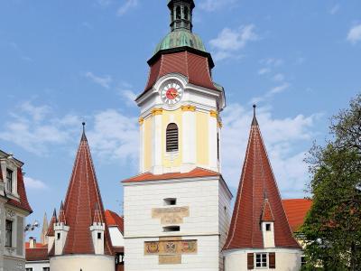 A large, white stone gatehouse with a central clock tower topped by a green dome and spire, flanked by two smaller round towers with red conical roofs, set against a blue sky with flowers in the foreground.
