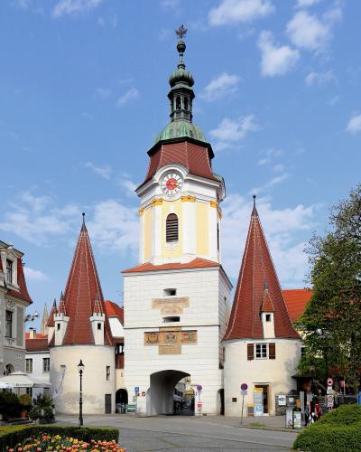 A large, white stone gatehouse with a central clock tower topped by a green dome and spire, flanked by two smaller round towers with red conical roofs, set against a blue sky with flowers in the foreground.