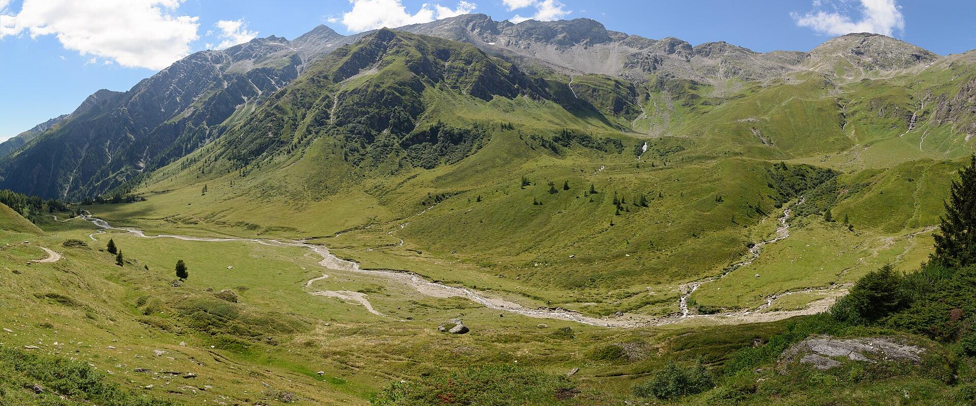 A wide alpine valley with a braided stream, green slopes, and rugged mountain peaks under a blue sky; no visible signage or structures.