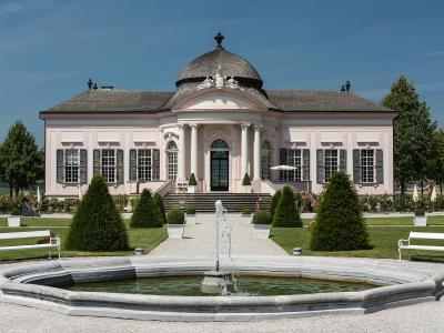 A pink Baroque pavilion with a central dome and columns, surrounded by a formal garden with a fountain and white benches.
