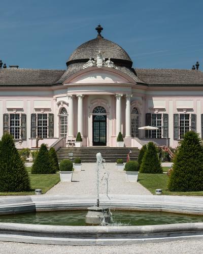 A pink Baroque pavilion with a central dome and columns, surrounded by a formal garden with a fountain and white benches.