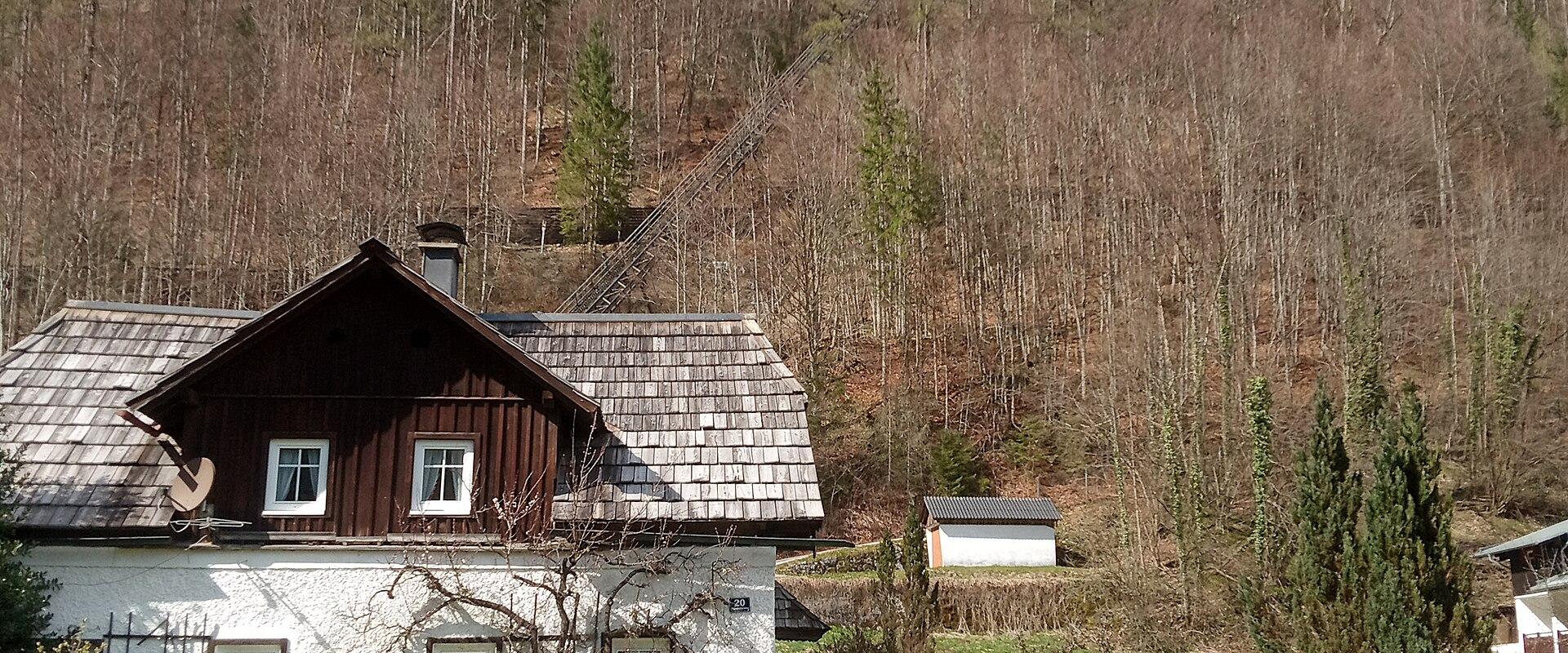 A traditional Austrian-style house with a white stucco lower level and dark wood upper story sits in the foreground, while a long, steep funicular railway ascends the forested mountainside behind it under a partly cloudy sky.