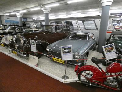 An indoor museum exhibit displaying vintage automobiles and a red motorcycle, with informational placards and a yellow license plate reading "MUSEUM-KAPRUN" on the front of a silver car.
