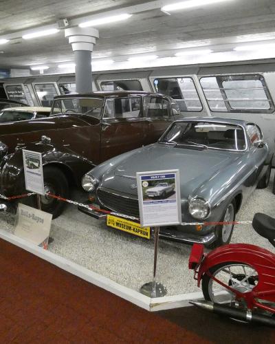 An indoor museum exhibit displaying vintage automobiles and a red motorcycle, with informational placards and a yellow license plate reading "MUSEUM-KAPRUN" on the front of a silver car.