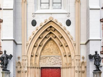 A close-up view of the ornate Gothic main portal of a large white stone church, featuring a red door, sculpted tympanum, and flanking statues.