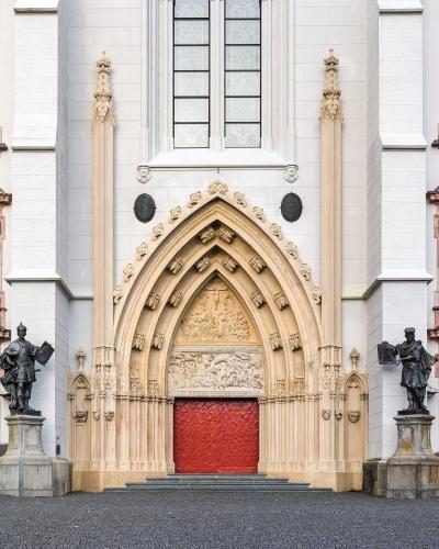 A close-up view of the ornate Gothic main portal of a large white stone church, featuring a red door, sculpted tympanum, and flanking statues.
