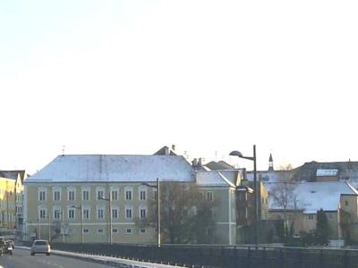 A street-level view of a town with a tall church tower on the right and a yellow road sign on the left indicating "Braunau" with an arrow pointing straight ahead.