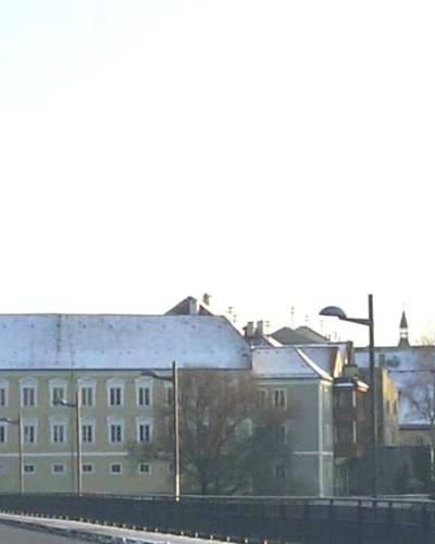 A street-level view of a town with a tall church tower on the right and a yellow road sign on the left indicating "Braunau" with an arrow pointing straight ahead.