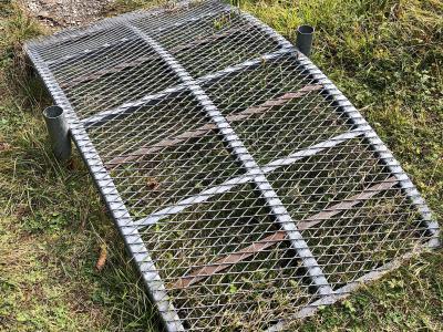 A close-up view of a rectangular metal grating bridge or walkway section lying on a grassy bank next to a dirt path.