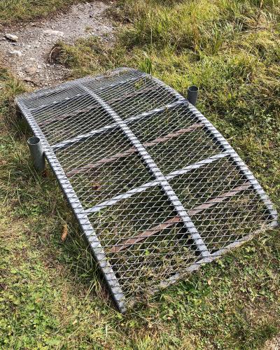 A close-up view of a rectangular metal grating bridge or walkway section lying on a grassy bank next to a dirt path.